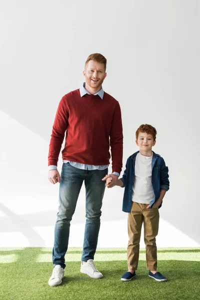 Вид в полный рост счастливых рыжих отца и сына, держащихся за руки и улыбающихся камере на сером фоне — стоковое фото