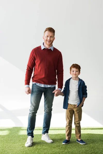 Vista completa de feliz pelirroja padre e hijo tomados de la mano y sonriendo a la cámara en gris - foto de stock