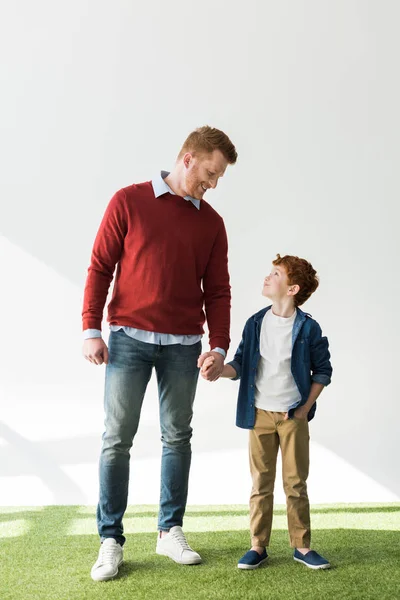 Glückliche rothaarige Vater und Sohn Händchen haltend und einander lächelnd auf grau — Stockfoto