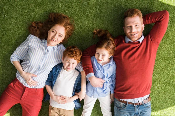 Draufsicht auf eine glückliche rothaarige Familie mit zwei Kindern, die zusammen im Gras liegen und in die Kamera lächeln — Stockfoto