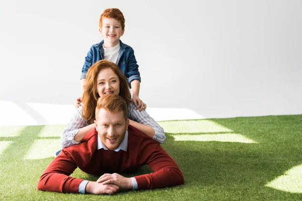 Feliz pelirroja familia con un niño acostado juntos en la hierba y sonriendo a la cámara en gris - foto de stock