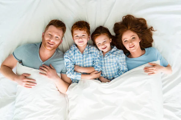 Felice rossa famiglia in pigiama sdraiato a letto e sorridente alla macchina fotografica — Foto stock