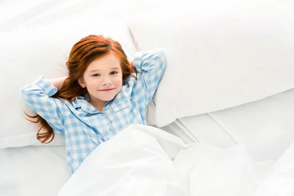 Adorable niño pequeño en pijama acostado en la cama y sonriendo a la cámara - foto de stock