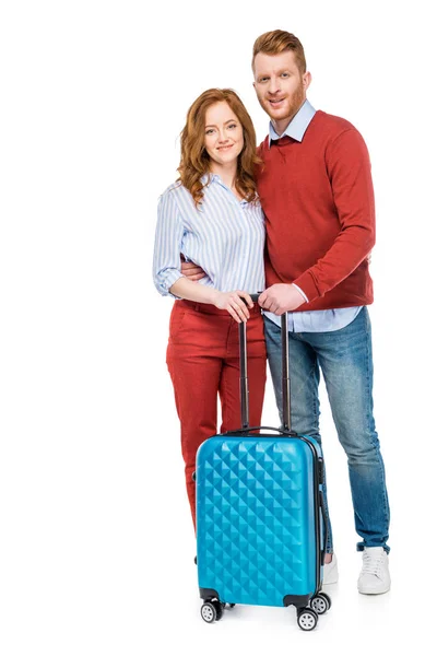 Счастливая рыжая пара, стоящая с чемоданом и улыбающаяся в камеру, изолированную на белом — стоковое фото