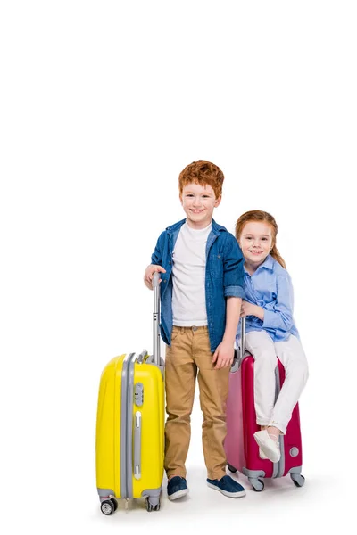Прелестные счастливые рыжие дети с чемоданами, улыбающиеся в камеру изолированы на белом — стоковое фото