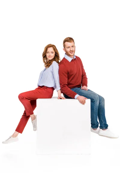 Full length view of happy redhead couple sitting on white cube and smiling at camera isolated on white — Stock Photo