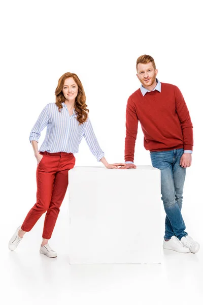 Visão comprimento total do casal ruiva feliz inclinando-se no cubo branco e sorrindo para a câmera isolada no branco — Fotografia de Stock