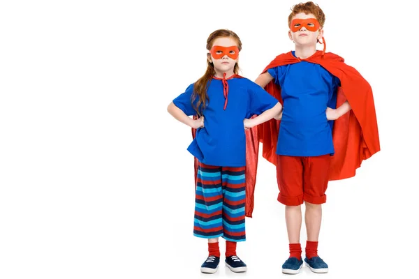 Super niños en máscaras y capas de pie con las manos en la cintura y mirando a la cámara aislada en blanco - foto de stock