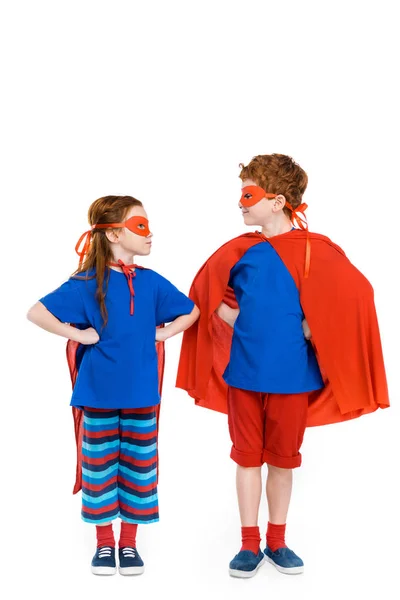 Super kids in masks and cloaks standing with hands on waist and looking at each other isolated on white — Stock Photo