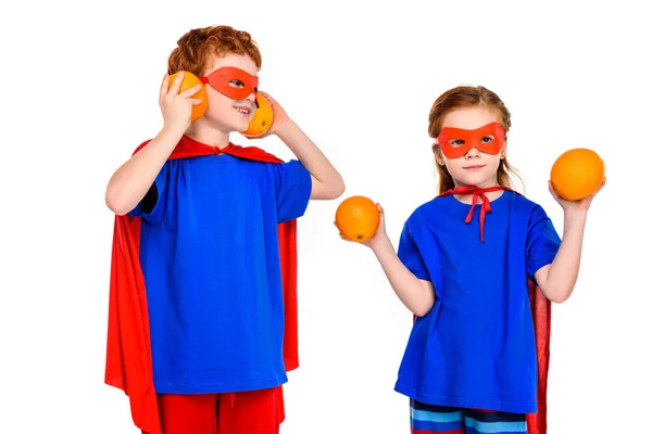 Lindos super niños en máscaras y capas que sostienen naranjas aisladas en blanco — Stock Photo