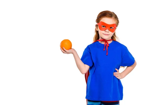 Super chico en máscara y capa sosteniendo naranja y sonriendo a la cámara aislado en blanco - foto de stock