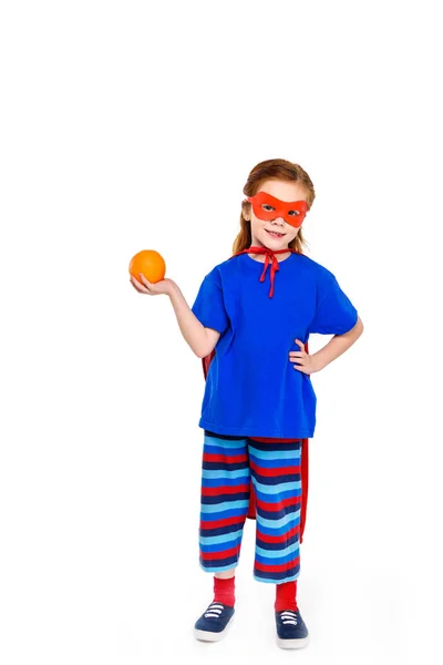 Adorable child in superhero costume holding orange and smiling at camera isolated on white — Stock Photo