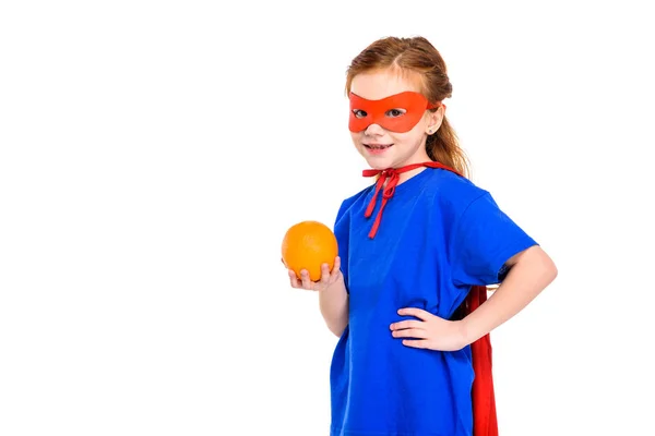 Super niño en máscara y capa sosteniendo naranja y sonriendo a la cámara aislada en blanco - foto de stock