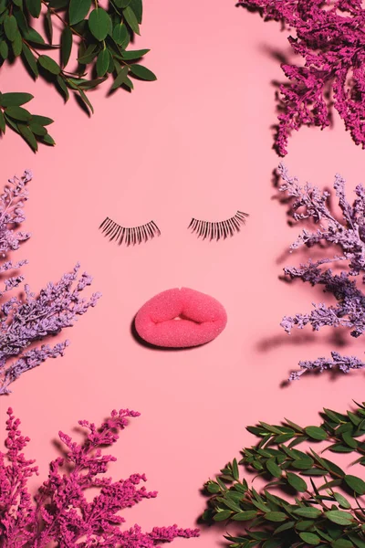 Top view of face made of sponge and false eyelashes surrounded with flowers on pink surface — Stock Photo