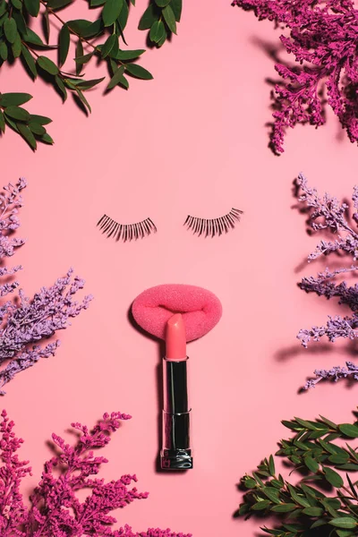 Vista superior de la cara hecha de esponja con pestañas postizas y lápiz labial rodeada de flores en la superficie rosa - foto de stock