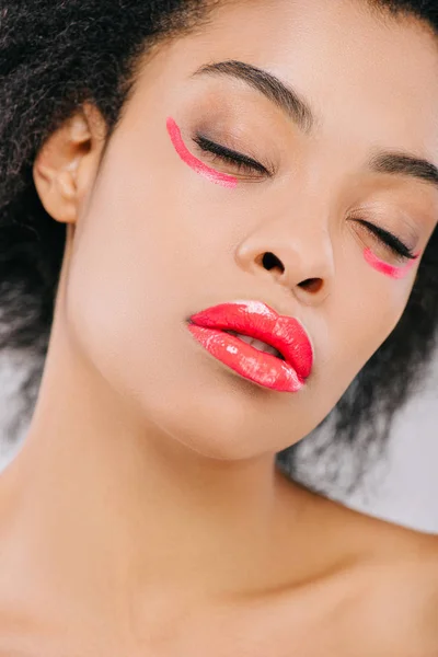 Sensual joven con labios rojos y pinceladas rojas bajo ojos cerrados aislados en gris - foto de stock