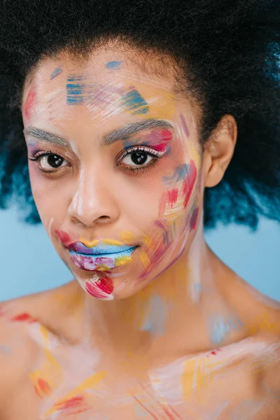 Jeune femme afro-américaine avec des coups de peinture sur le visage en regardant la caméra isolée sur bleu — Photo de stock