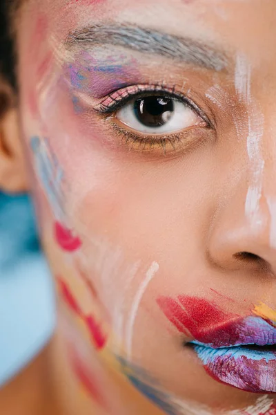 Plan recadré de jeune femme avec des coups colorés sur le visage en regardant la caméra sur bleu — Photo de stock