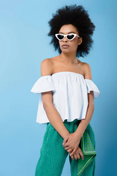 Mujer afroamericana joven en elegante parte superior fuera del hombro y gafas de sol aisladas en azul - foto de stock