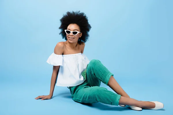 Jeune femme afro-américaine dans des vêtements élégants et des lunettes de soleil assis sur le sol bleu — Photo de stock