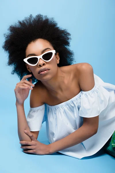 Jeune femme afro-américaine dans des vêtements élégants et des lunettes de soleil couché sur le sol bleu — Photo de stock