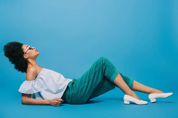 Attraktive junge Frau in stylischer Kleidung und Sonnenbrille auf dem Boden liegend auf blau — Stockfoto