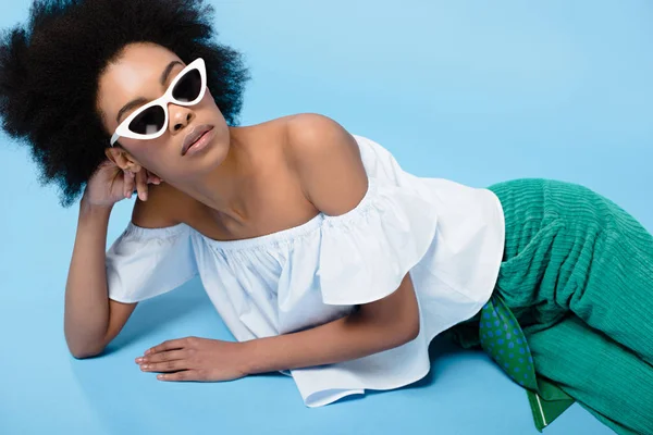 Belle jeune femme dans des vêtements élégants et des lunettes de soleil couché sur le sol sur bleu — Photo de stock