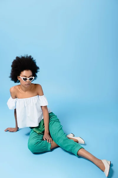 Beautiful young woman in stylish clothes and sunglasses sitting on floor on blue — Stock Photo