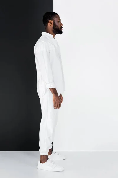 Vue latérale de l'homme afro-américain élégant debout dans des vêtements blancs près du mur noir et blanc — Photo de stock