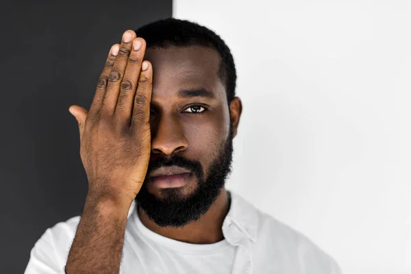 Bel homme afro-américain élégant en vêtements blancs fermant les yeux avec la main et regardant la caméra — Photo de stock