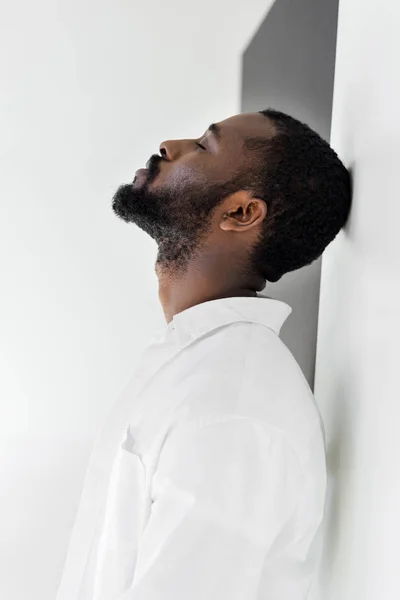 Seitenansicht eines hübschen, stilvollen afrikanisch-amerikanischen Mannes in weißer Kleidung, der sich mit geschlossenen Augen an die Wand lehnt — Stockfoto