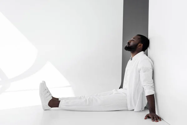 Vue latérale du bel homme afro-américain élégant assis sur le sol et appuyé sur le mur en vêtements blancs — Photo de stock