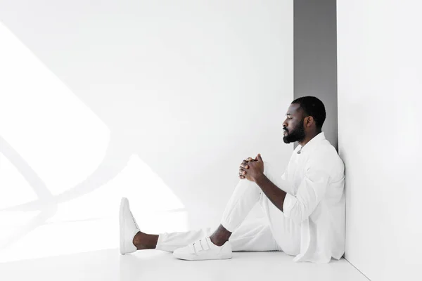 Side view of handsome stylish african american man sitting on floor in white clothes — Stock Photo