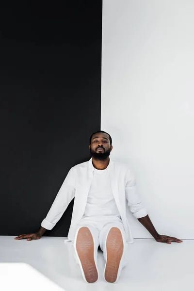 Bel homme afro-américain élégant assis sur le sol près du mur noir et blanc et regardant la caméra — Photo de stock