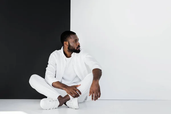 Красивий стильний афроамериканський чоловік сидить на підлозі біля чорно-білої стіни і дивиться в сторону — стокове фото