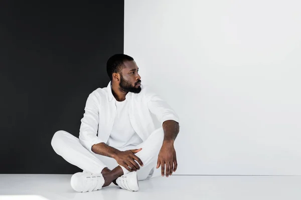 Bel homme afro-américain élégant assis sur le sol près du mur noir et blanc et regardant loin — Photo de stock