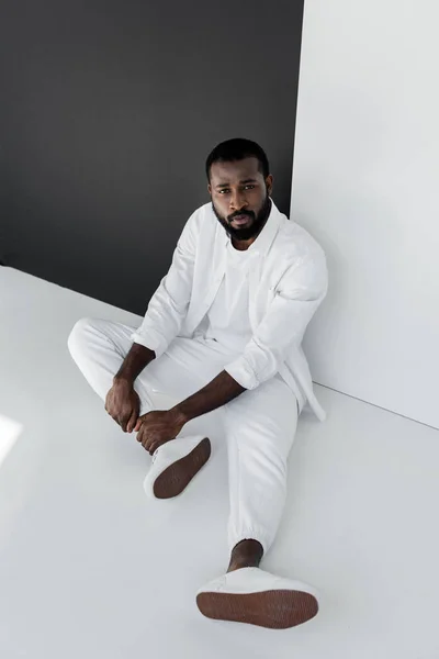 Vista de alto ângulo de belo homem americano africano elegante sentado no chão perto de parede preta e branca — Fotografia de Stock