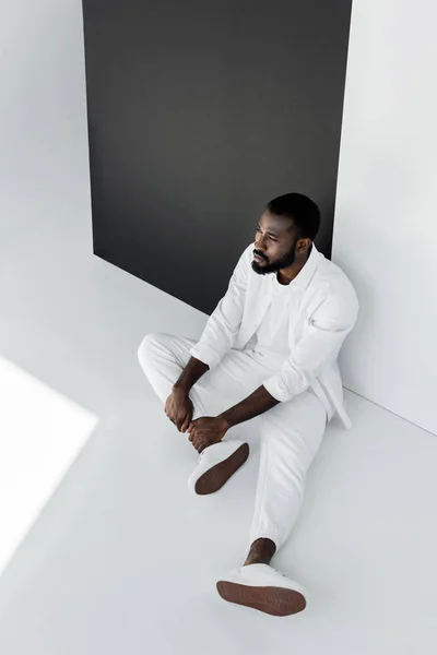 Vue grand angle de bel homme afro-américain élégant assis sur le sol en vêtements blancs — Photo de stock