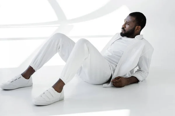 Bonito elegante afro-americano homem em roupas brancas deitado no branco — Fotografia de Stock