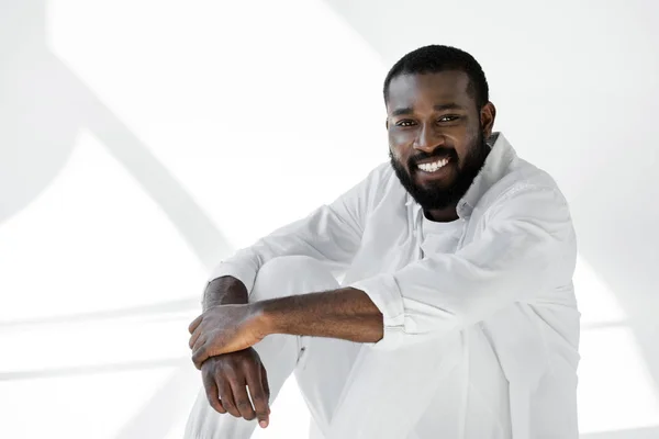 Smiling handsome african american man in white clothes sitting and looking at camera on white — Stock Photo