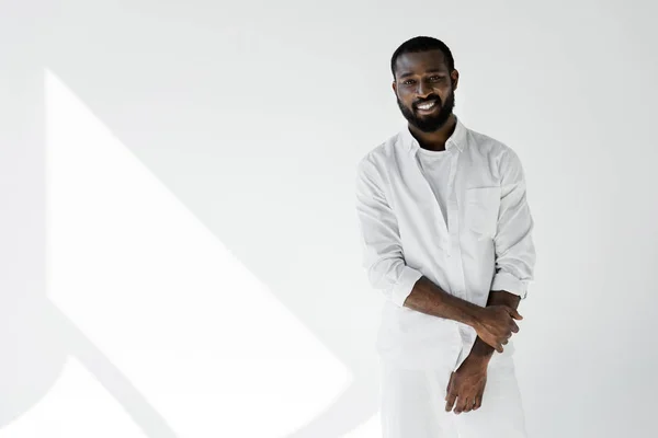 Улыбающийся стильный африканский американец в белой одежде смотрит в камеру на белом — стоковое фото