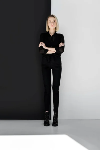 Attraktive, stylische blonde Frau in schwarzer Kleidung, die mit verschränkten Armen nahe der schwarz-weißen Mauer steht — Stockfoto