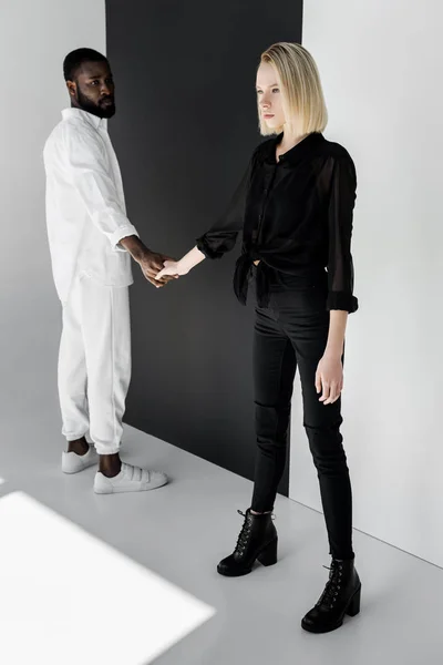 Pareja multicultural cogida de la mano cerca de la pared en blanco y negro - foto de stock