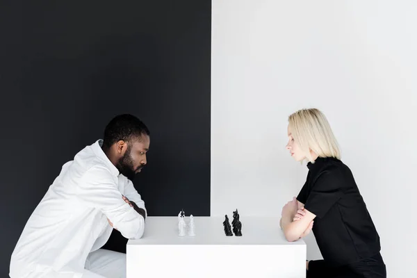 Vue latérale du petit ami afro-américain et petite amie blonde jouant aux échecs près du mur noir et blanc — Photo de stock