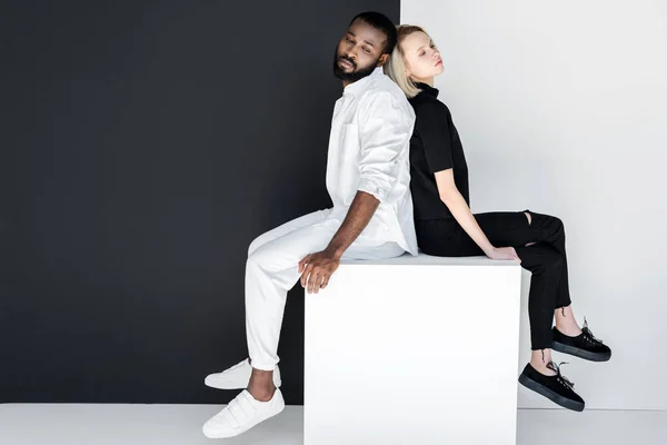 African american boyfriend and blonde girlfriend sitting back to back on white cube — Stock Photo