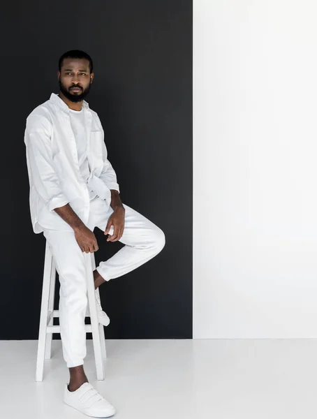 Bel homme élégant afro-américain en vêtements blancs assis sur une chaise près du mur noir et blanc — Photo de stock