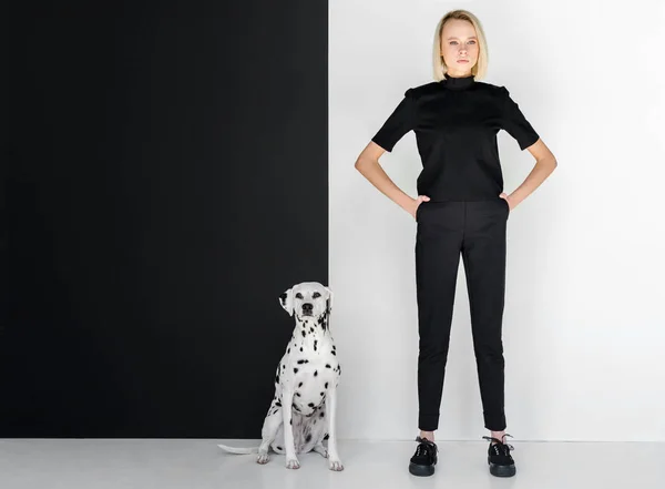 Beautiful stylish blonde woman in black clothes standing near black and white wall with dalmatian dog — Stock Photo