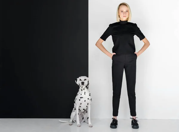 Belle femme blonde élégante en vêtements noirs debout près du mur noir et blanc avec chien dalmate — Photo de stock