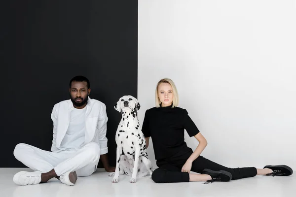 Casal multicultural sentado perto de parede preto e branco com cão dalmatian — Fotografia de Stock