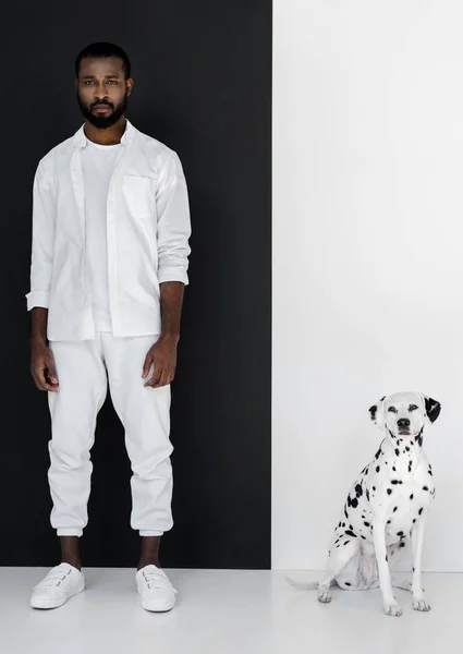 Bel homme afro-américain élégant en vêtements blancs avec chien dalmate près du mur noir et blanc — Photo de stock